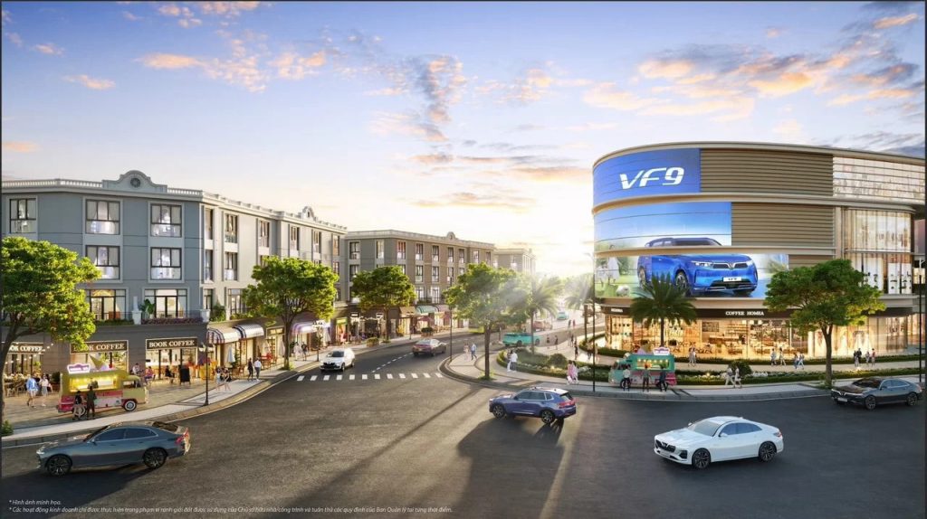 Vinhomes Hòa Long Bắc Ninh 12 Phối cảnh khu thương mại tại Vincom Mega Mall Vinhomes Hòa Long - Bắc Ninh, với các tòa nhà hiện đại, cửa hàng, quán cà phê, và biển quảng cáo xe VinFast VF 9, cùng không gian đường phố nhộn nhịp có xe cộ và cư dân qua lại.